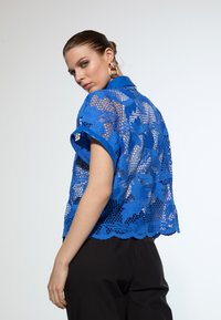 Blue lace short-sleeve shirt featuring floral patterns, a scalloped hem, and a collared neckline, paired with black trousers.