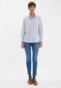 Chemise bleu clair à manches longues, avec une seule poche poitrine ; portée avec un jean bleu et des chaussures brunes avec un accent en nœud.