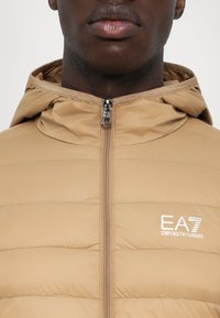 Prim-plan cu o persoană îmbrăcată într-o jachetă de tip puffer bej, cu glugă și fermoar frontal, având pe piept logo-ul alb EA7 Emporio Armani.