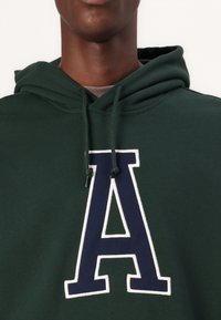 Grønn hettegenser med et stort, hvitkantet marineblått "A"-logo på forsiden. Har snor i hetten og en myk tekstur.
