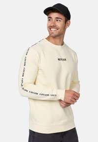 Sudadera crema de manga larga, con un logotipo de texto negro en la parte frontal y a lo largo de las mangas, puños y dobladillo acanalados, fabricada con una suave mezcla de algodón.