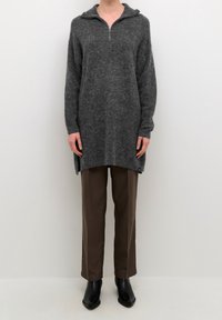 Grauer Strick-Tunika mit einem Viertel-Reißverschluss-Kragen, seitlichen Schlitzen und langen Ärmeln, kombiniert mit braunen Hosen und schwarzen Ankle-Boots.