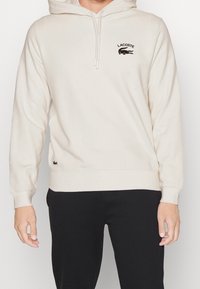 Sweat à capuche beige clair en tissu doux, doté d'une poche kangourou à l'avant, d'une capuche avec cordon de serrage, et d'un logo noir Lacoste sur la poitrine.