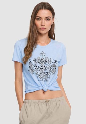 Giovane donna con capelli lunghi che indossa una T-shirt azzurra legata con scritte a motivo e pantaloni beige, su sfondo semplice.