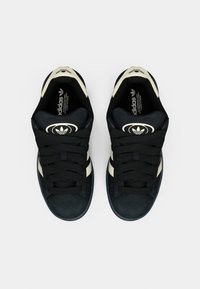 Melnas zamšādas sporta kurpes ar krēmkrāsas svītrām, apaļu logotipu un bieziem melniem šņorēm. Gluda tekstūra, zems siluets un mīkstināta zolīte.