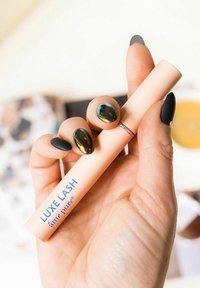 Luxe Lash mascara röret hålls i en hand; röret är mjuk rosa med blå text; glansiga, mörka naglar; slät textur, cylindrisk form.