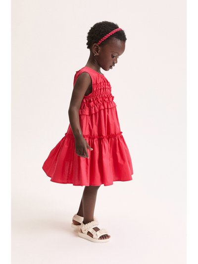 Jeune fille portant une robe rouge à volants et des sandales blanches ornées de perles, regardant vers le bas avec un bandeau rouge dans ses courts cheveux bouclés.