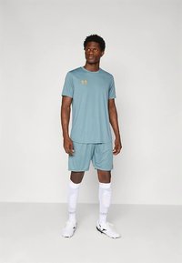 Sportovec v světle modrém tričku odvádějícím vlhkost a odpovídajících šortkách, s bílými podkolenkami a bílými sportovními botami. Zlatý detail loga.