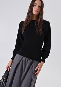 Giovane donna con lunghi capelli castani che indossa un top nero a maniche lunghe e una gonna grigia, mentre tiene una borsa nera su uno sfondo neutro.