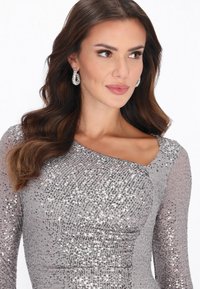 Silbernes Paillettenkleid mit asymmetrischem One-Shoulder-Design und strukturierten langen Ärmeln. Kombiniert mit funkelnden Tropfenohrringen.