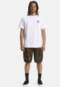 Bílá bavlněná tričko s logem na levé hrudi, spárované s maskáčovými cargo šortkami s kapsami. Černé ponožky a tenisky dotvářejí celý outfit.