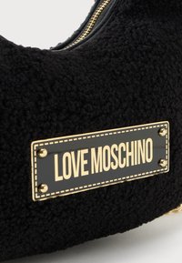 Melnā teksturētā soma ar zelta toni rāvējslēdzēju un taisnstūrveida melnu un zelta etiķeti ar uzrakstu "LOVE MOSCHINO", pievienota ar četriem zelta kniedēm.