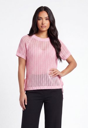 Kvinde med langt mørkt hår iført en lyserød kortærmet strikket sweater og sorte bukser, stående med den ene hånd på hoften.