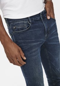 Sötétkék farmernadrág, slim fit szabással, látható varrással, klasszikus öt zsebes kialakítással és diszkrét kopási részletekkel.