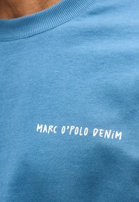 Μπλε βαμβακερό πουκάμισο με λευκό κείμενο "MARC O'POLO DENIM" τυπωμένο στο στήθος κοντά στον γιακά.