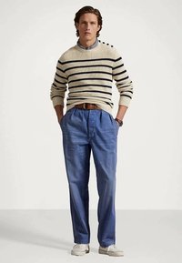 Ung mann står med hendene i lommene, iført en kremfarget og marineblå stripete genser, blå høytalende plisserte bukser og hvite sko.