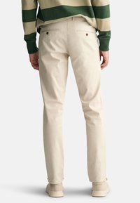 Beige cotton pants with a slim fit, featuring two back pockets with button closures and a smooth texture. Worn with beige footwear.