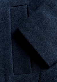 La tela de lana de color azul marino presenta una superficie suave y texturizada con un detalle de parche cuadrado. El diseño es limpio y minimalista.