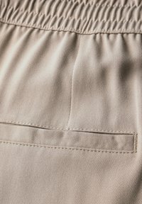 Tissu en coton beige avec une texture lisse, doté d'une taille élastique et d'une poche arrière avec un contour piqué pour une touche de détail supplémentaire.