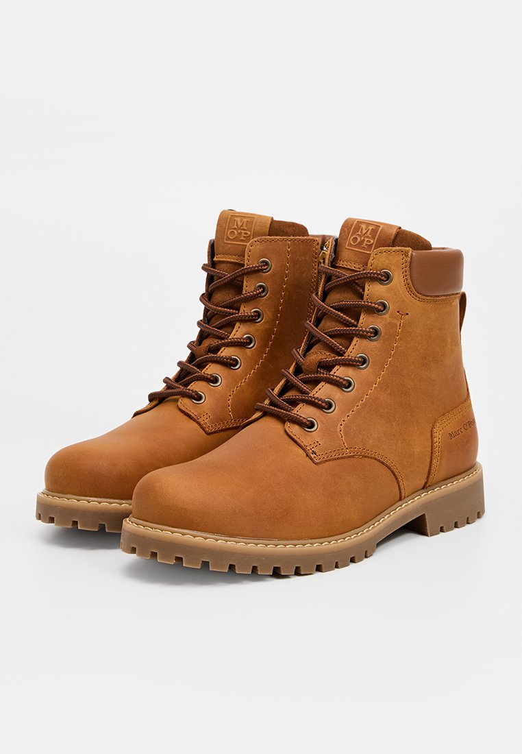 Bottines chelsea en cuir brun à finition lisse, toe rond, œillets en métal et semelle en caoutchouc robuste. Présente des lacets marron et un détail de logo sur le côté.