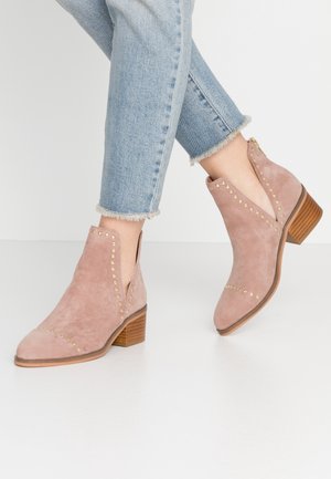 Pudrowo-różowe zamszowe botki za kostkę z bocznymi wycięciami, złotymi ćwiekami i zakrytym drewnianym obcasem. Noszone z dżinsami o postrzępionych końcówkach.