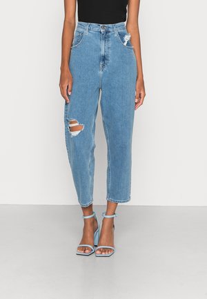 Vrouw draagt een hoge taille, lichtblauwe gescheurde spijkerbroek met een zwart mouwloos topje en lichtblauwe sandalen met open teen en blokhak.