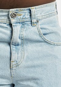 Light blue denim jeans feature a high waist, contrasting yellow stitching, and a metal button with a skull design. Subtle texture detail visible.