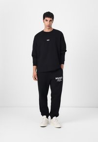 Black sweatshirt and sweatpants with white logo detailing, made from cotton fabric. Model wearing white sneakers, standing against a plain background.
