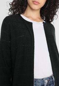 Kvinna med lockigt hår som bär en svart öppen cardigan över en vit t-shirt och blåjeans, visad från axlarna till midjan.