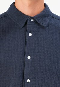 Donkerblauwe shirt van een gestructureerde, geweven stof, met een puntkraag en witte knopen langs de voorste plaket.