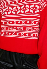 Punainen neulottu neule valkoisilla geometrisilla kuvioilla ja etupuolella jouluisella tekstillä "NOËL JOYEUX NOËL", rennolla istuvuudella ja teksturoidulla kankaalla.