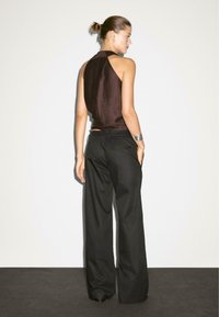 Une femme se tient de dos face à la caméra, portant un haut sans manches sombre et un pantalon large noir, devant un arrière-plan neutre.