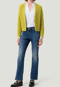 Helle gelbe Strickjacke über einem weißen Hemd mit Kragen, kombiniert mit blauen Schlagjeans und schwarzen Slippern mit silbernen Akzenten.