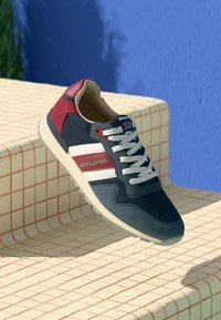 Mørkeblå og rød sneaker med hvide striber, øvre del i stof, polstret krave og gummisål, præsenteret på en gittermønstret overflade.