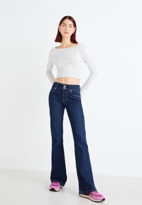 Haut court blanc à côtes avec ourlet festonné, associé à un jeans évasé bleu foncé avec deux poches avant et fermeture à bouton. Baskets roses.