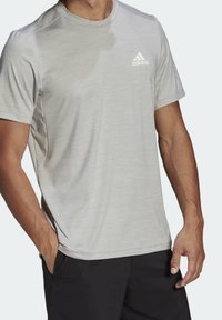 Ljusgrå atletisk T-shirt gjord av fukttransporterande tyg. Har rund hals, korta ärmar och en liten vit Adidas-logotyp på bröstet.