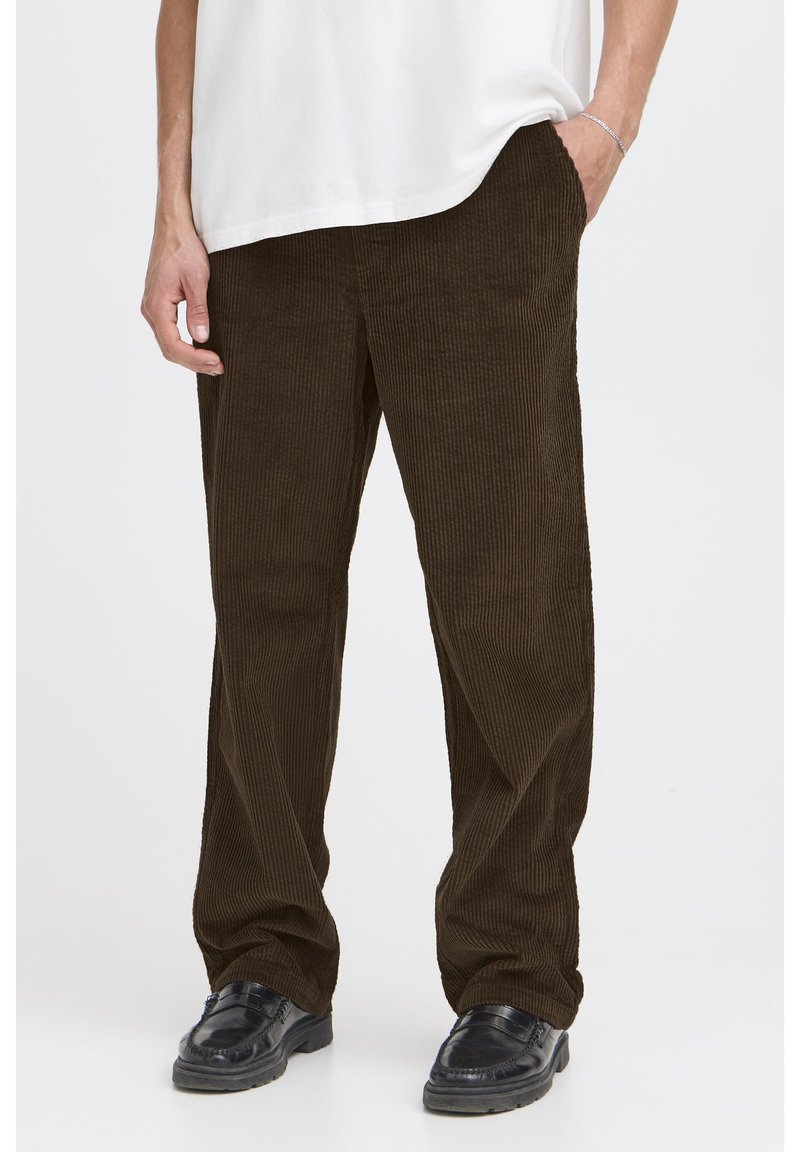 Brown corduroy trousers with a ribbed texture, featuring a loose fit, side pockets, and paired with black shoes. Simple white shirt above.