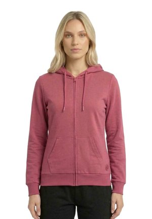 Une femme porte un sweat à capuche rose poudré zippé, avec un cordon de serrage à la capuche et des poches à l’avant, sur fond blanc.