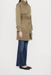 Long manteau trench couleur kaki avec une taille ceinturée, des boutons-pression et des poches latérales, assorti à un jean bleu évasé et des ballerines noires.
