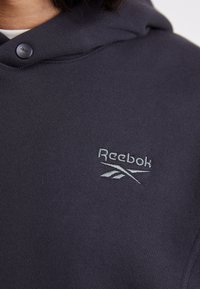 Felpa con cappuccio blu navy dal tessuto liscio, dotata di un bottone frontale e logo Reebok ricamato in grigio chiaro.