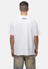 Witte katoenen T-shirt met korte mouwen, met een rond logo op het achterste nekgedeelte. De pasvorm is ontspannen en de stof lijkt glad.