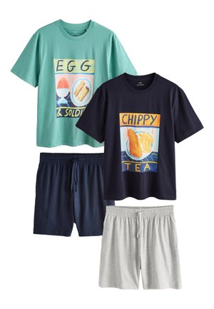 Mężczyzna o ciemnych włosach z tatuażem na lewej ręce stoi boso, ubrany w granatowy t-shirt z grafiką fish and chips oraz białe szorty, w sypialni.