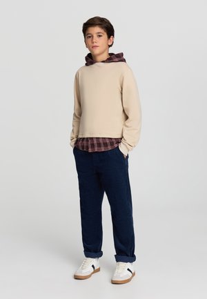 Felpa beige con cappuccio sopra una camicia a quadri bordeaux, abbinata a pantaloni in velluto a coste blu marina e sneaker bianche con strisce nere.