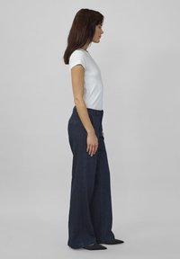 Zijprofiel van een persoon in een strak wit T-shirt en donkere wijde jeans, met lang donker haar, tegen een neutrale achtergrond.