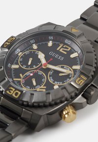 Reloj de acero inoxidable negro con acentos dorados, grandes marcadores numéricos, subesferas de cronógrafo, esfera texturizada y bisel rotatorio con marcadores de minutos.