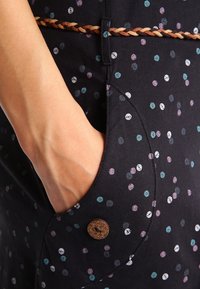 Zwarte stof met kleurrijke stippen. Bevat een gevlochten riem en een decoratieve knop op de zak, die de textuur en details benadrukken.