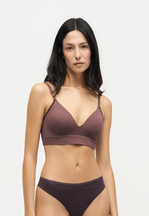 Femme aux longs cheveux foncés portant un soutien-gorge marron assorti et des sous-vêtements, debout devant un fond clair uni, faisant face vers l'avant.