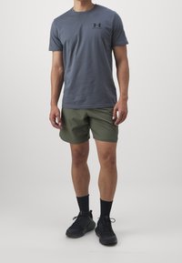 Camiseta de algodón gris con un logo negro, combinada con pantalones cortos verdes oliva. Zapatillas negras con calcetines negros completan el atuendo.