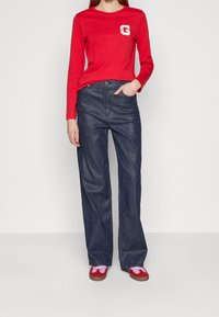 Rode longsleeve t-shirt met een witte letter "G" op de borst, gecombineerd met losse blauwe denim jeans en rode schoenen met roze accenten.