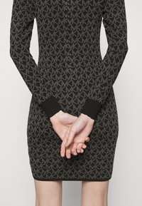 Robe en maille noire avec un motif de logo répété. Manches longues et poignets côtelés. Silhouette ajustée avec une longueur mi-cuisse.